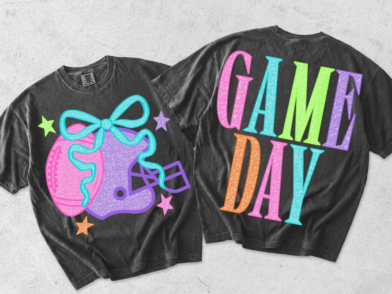 Two black graphic t-shirts displayed flat against a light gray textured background, showing the front and back designs. The front features a pink softball, purple football helmet with a turquoise bow, and colorful stars.