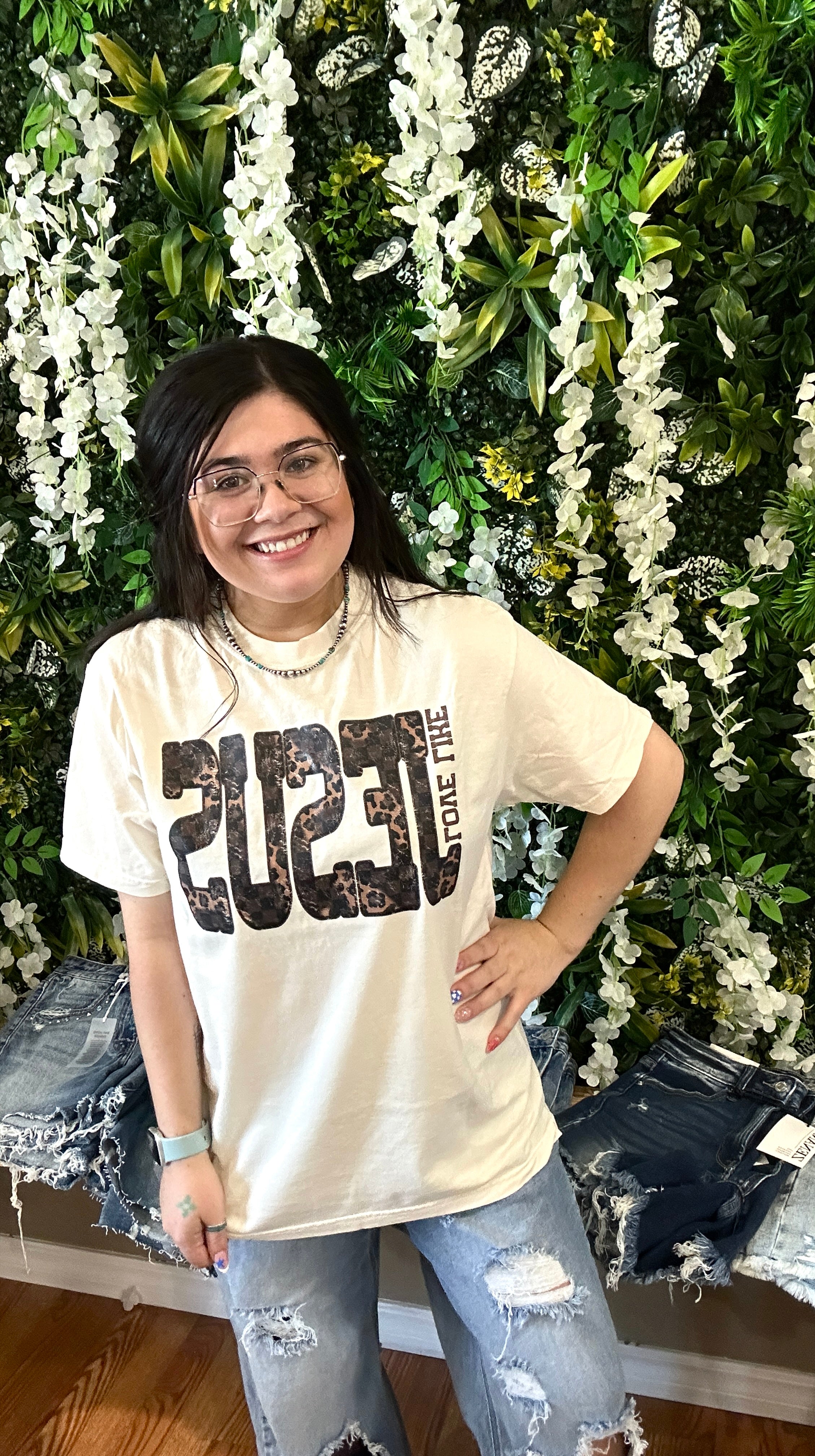 A woman wearing a cream-colored graphic t-shirt with distressed black and brown '2023' lettering, paired with ripped blue jeans, standing in front of a green foliage and white flower wall.