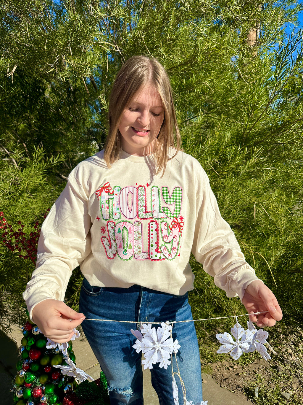 Person wearing a 'Holly Jolly' Long Sleeve T-Shirt outdoors with trees in the background