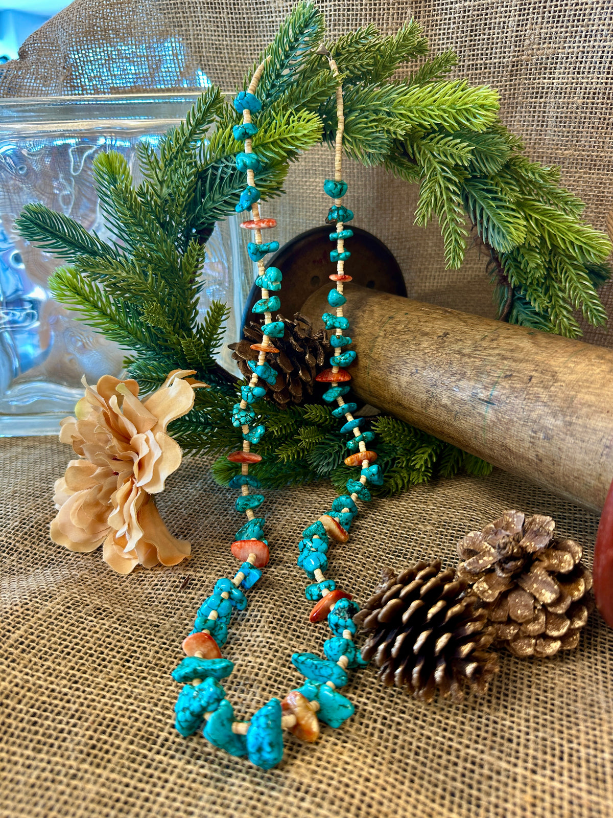 Turquoise beaded necklace with coral accents on a rustic background with pine branches and a pine cone.