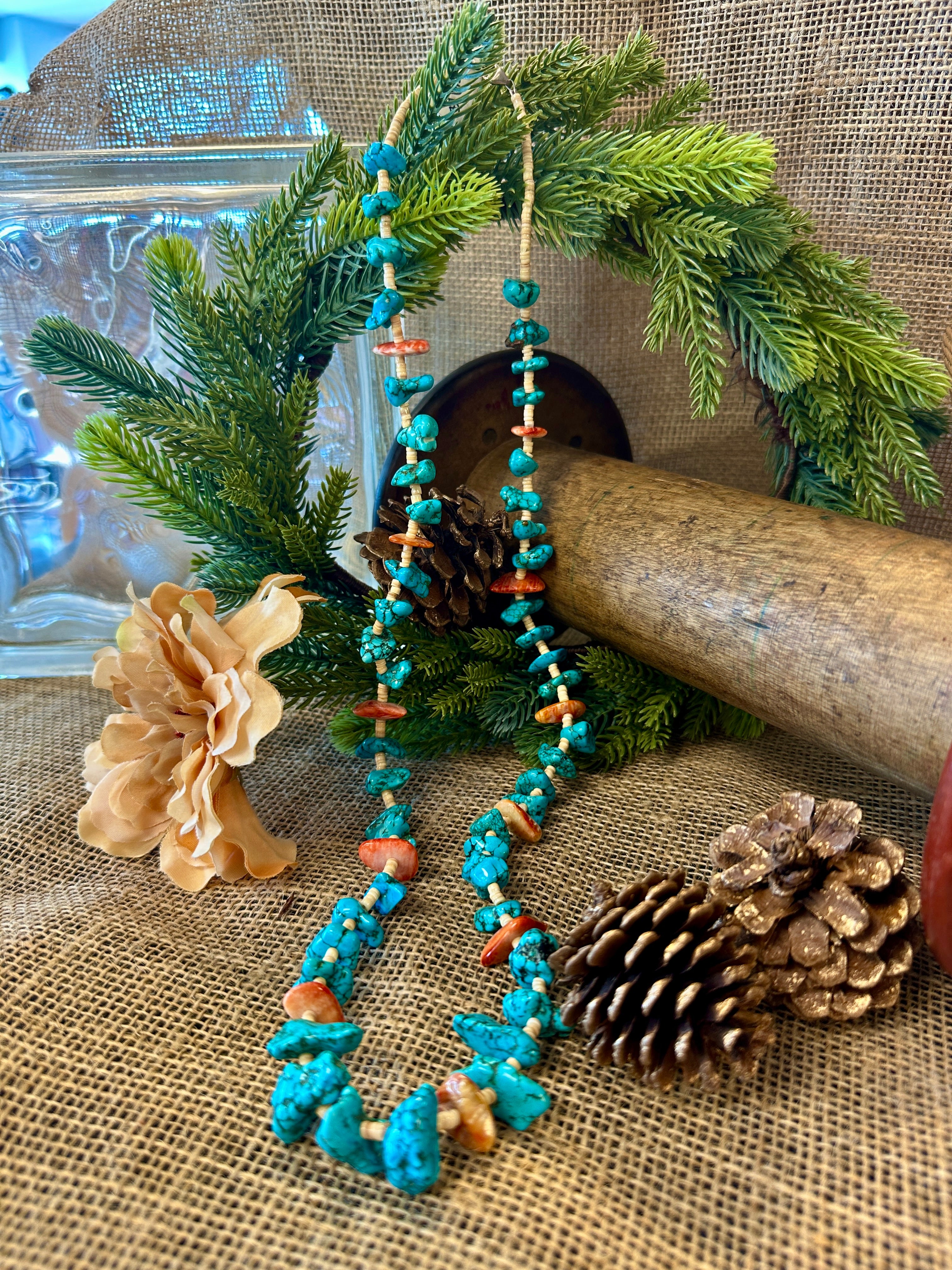 Turquoise beaded necklace with coral accents on a rustic background with pine branches and a pine cone.