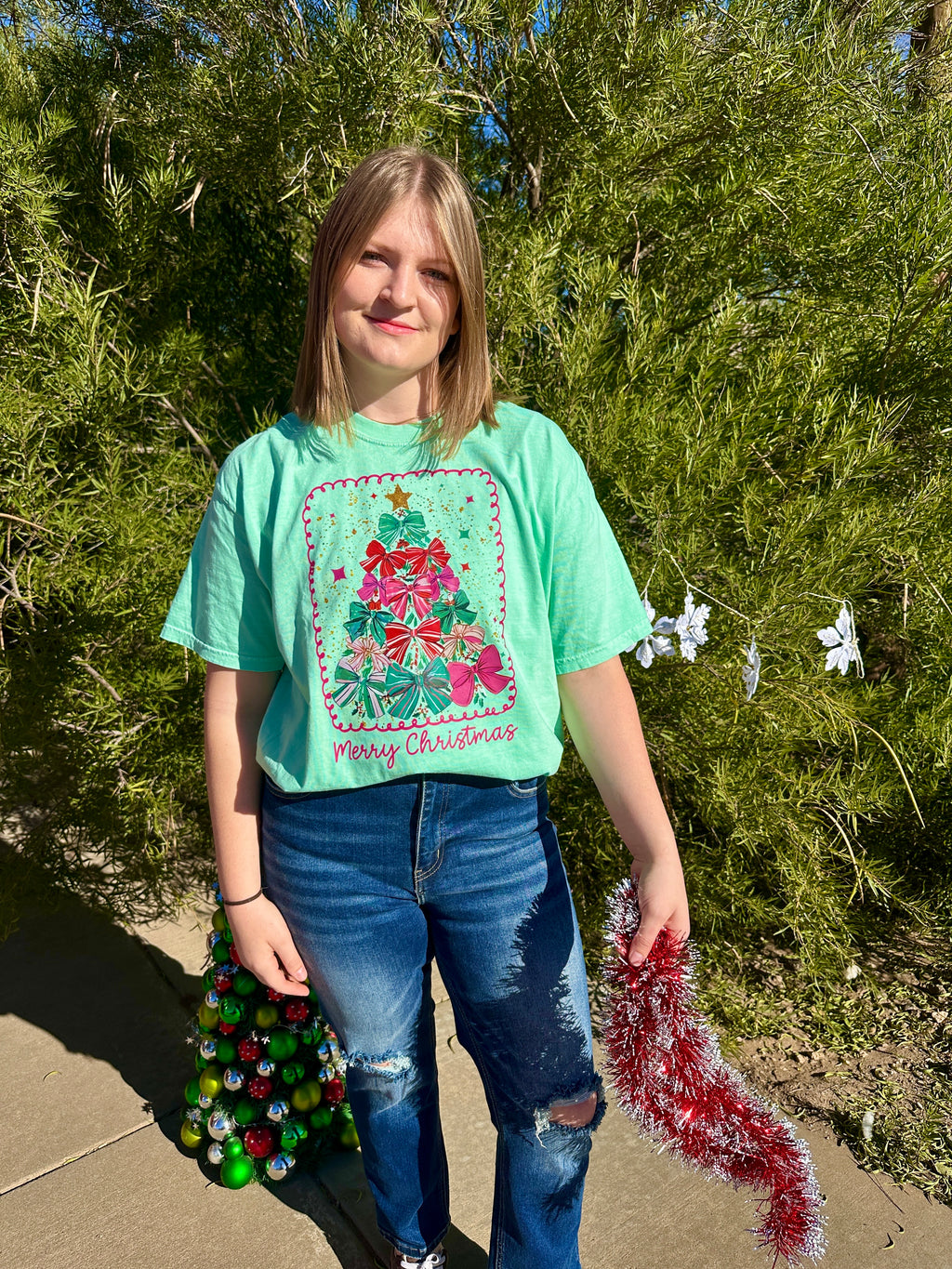 Person wearing a mint green Christmas-themed t-shirt holding festive decorations outdoors.