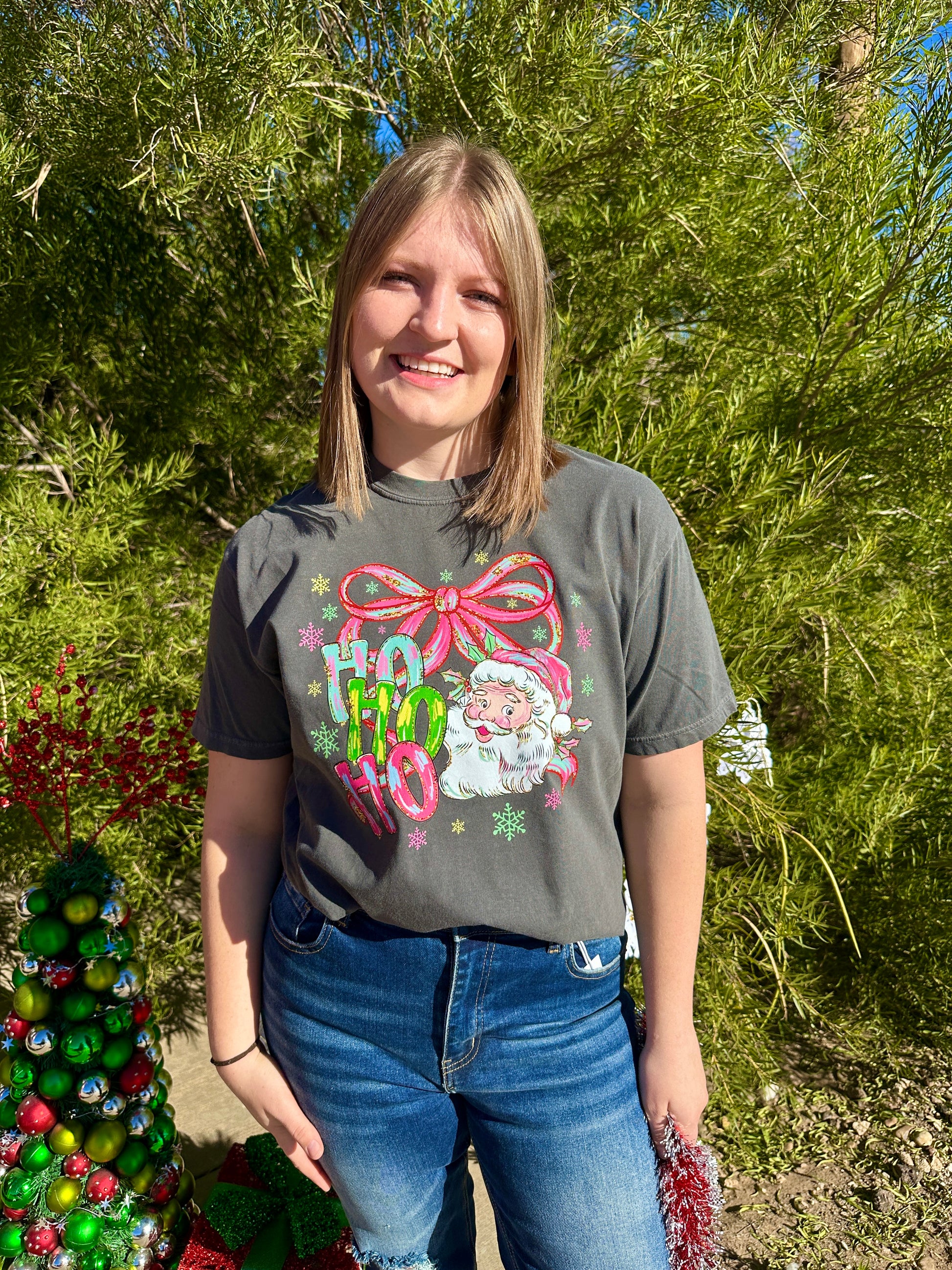 Person wearing a festive t-shirt with a pink bow and text, standing outdoors with greenery in the background.
