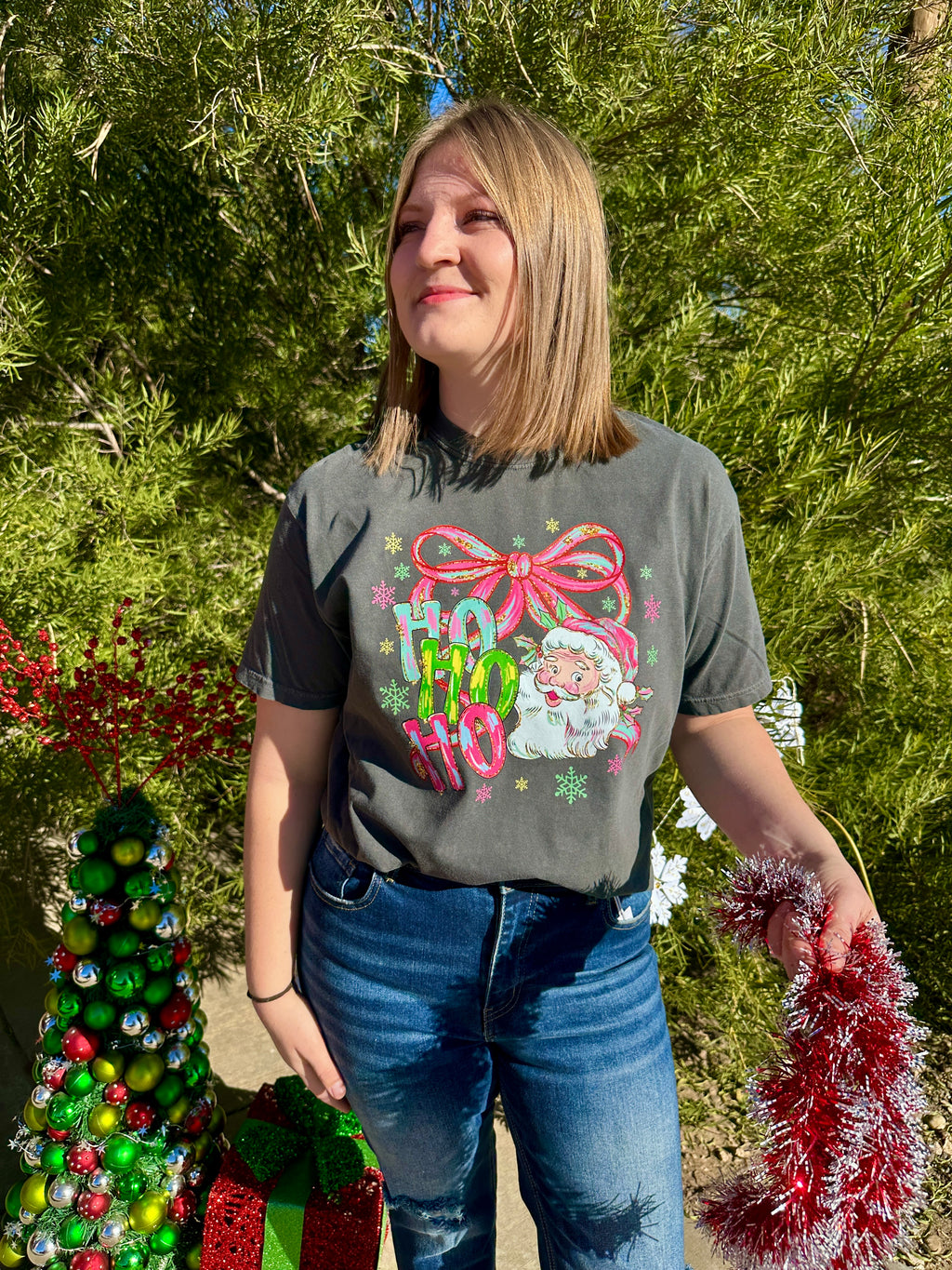 Person wearing a festive t-shirt with Christmas-themed graphics, standing outdoors with greenery and decorations.